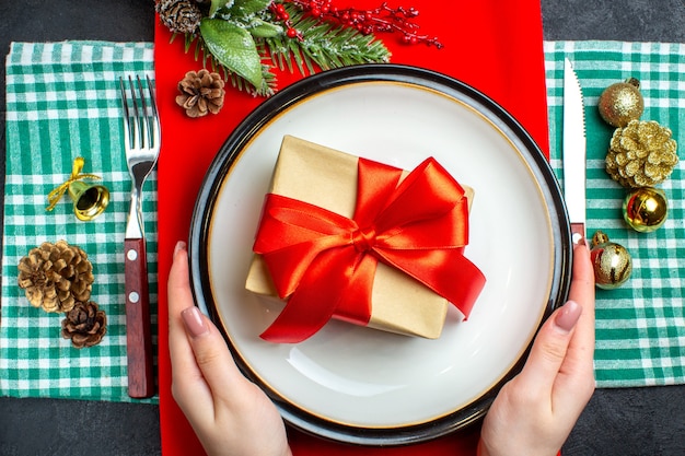 Bovenaanzicht van hand met een mooie geschenkdoos met boogvormig rood lint op een bord en bestek set decoratie-accessoires op groene gestripte handdoek