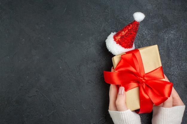 Bovenaanzicht van hand met mooi cadeau met boogvormig lint naast kerstman hoed op donkere achtergrond