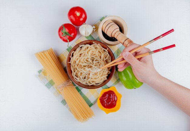 Bovenaanzicht van hand met stokjes en macaroni pasta in kom met tomaten zwarte peper ketchup knoflook peper en vermicelli op geruite doek en wit