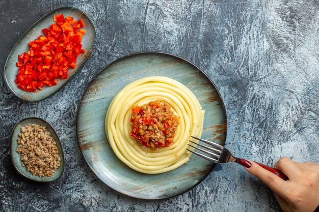 Bovenaanzicht van hand met vork op heerlijke pastamaaltijd op een blauw bord geserveerd met tomaat en vlees voor het diner naast de ingrediënten op ijsachtergrond