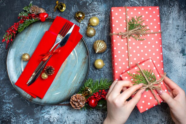 Bovenaanzicht van hand openen van een geschenkdoos en bestekset met rood lint op een decoratief servet op een blauw bord en kerstaccessoires en kerstsok op donkere achtergrond