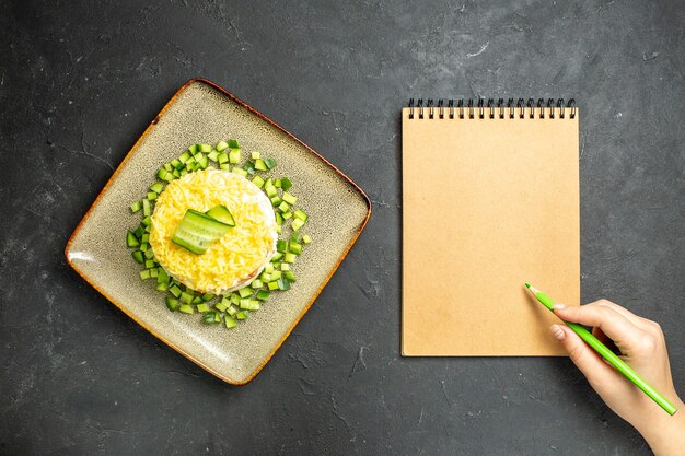 Bovenaanzicht van handschrift op notitieboekje en heerlijke salade geserveerd met gehakte komkommer op donkere achtergrond