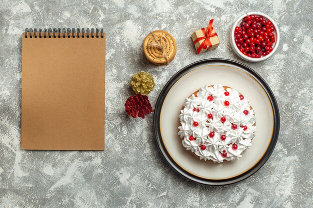 Bovenaanzicht van heerlijke cake met roombes op een bord en geschenkdozen gestapelde koekjes conifer kegels naast notebook op grijze achtergrond