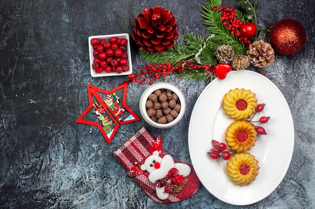 Gratis foto bovenaanzicht van heerlijke koekjes op een witte plaat en cornell-chocolade in kommen dennentakken aan de linkerkant op een donkere ondergrond