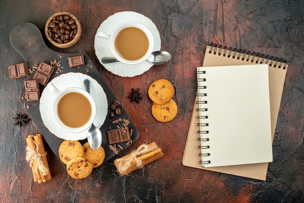 Bovenaanzicht van heerlijke koffie in witte kopjes op houten snijplank koekjes, kaneel limoenen, chocoladerepen, spiraalvormige notitieboekjes