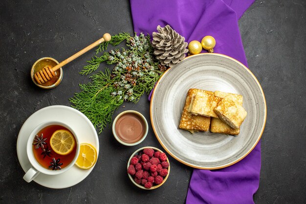 Bovenaanzicht van heerlijke pannenkoeken op een witte plaat chocolade en frambozen decoratie accessoires op paarse handdoek en een kopje zwarte thee op zwarte achtergrond