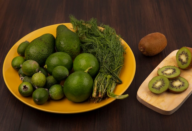 Bovenaanzicht van heerlijke plakjes kiwi op een houten keukenbord met avocado's feijoas en limoenen (lemmetjes) op een gele plaat op een houten muur