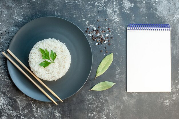 Bovenaanzicht van heerlijke rijstmaaltijd geserveerd met groen en eetstokjes op een zwarte plaat, paprika's laten een spiraalvormig notitieboekje op donkere achtergrond achter
