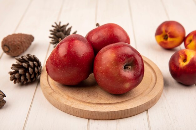 Bovenaanzicht van heerlijke rode appels op een houten keukenbord met perziken en dennenappels geïsoleerd op een wit houten oppervlak