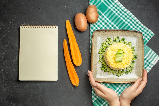 Bovenaanzicht van heerlijke salade geserveerd met gehakte komkommer op halfgevouwen groene gestripte handdoek en wortelaardappelen naast notitieboekje op donkere achtergrond