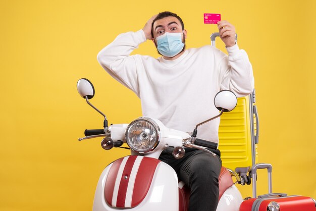 Bovenaanzicht van het reisconcept met een verwarde man met een medisch masker die op een motorfiets zit met een gele koffer erop en een bankkaart vasthoudt