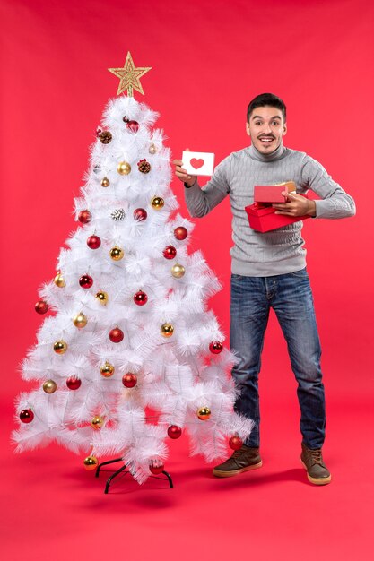 Bovenaanzicht van jonge volwassene in een grijze blouse die zich dichtbij de versierde witte kerstboom bevindt en zijn giften houdt