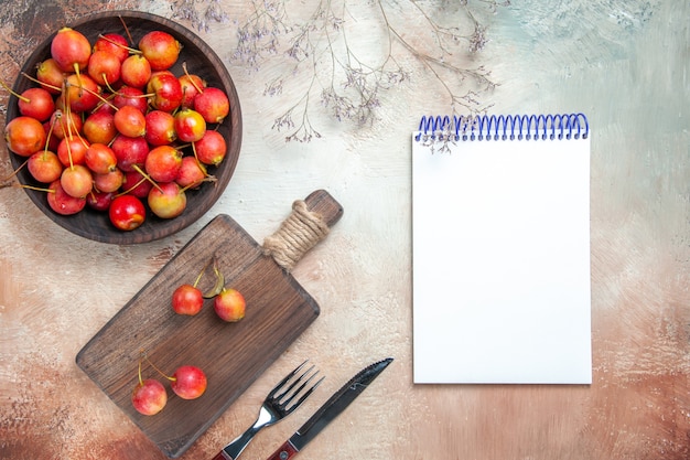 Bovenaanzicht van kersen bessen op het bord kom met kersen vork mes witte notebook