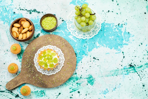 Bovenaanzicht van kleine cake met heerlijke room en gesneden en verse groene druiven cookies geïsoleerd op blauw, cake zoet fruit suiker