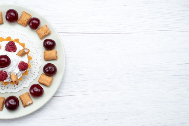 Bovenaanzicht van kleine romige cake met frambozen en kleine koekjes op wit