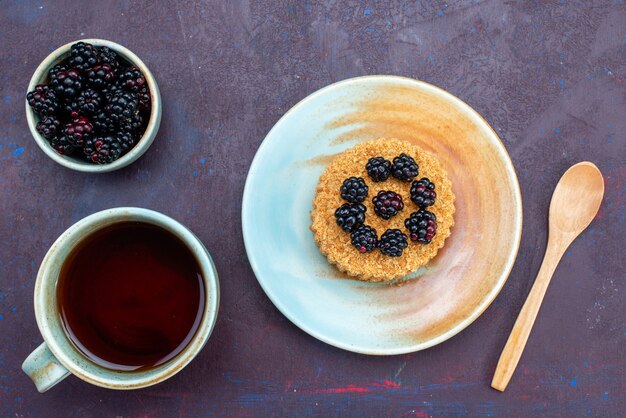 Bovenaanzicht van kleine ronde cake met bessen in plaat samen met kopje thee op donkere ondergrond