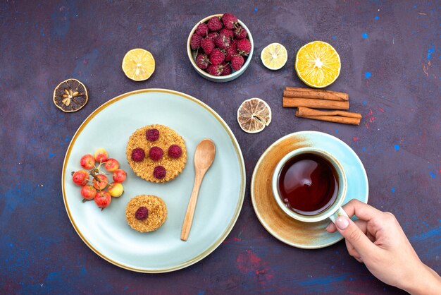 Bovenaanzicht van kleine ronde cake met verse frambozen in plaat met vruchten kaneelthee op het donkere oppervlak