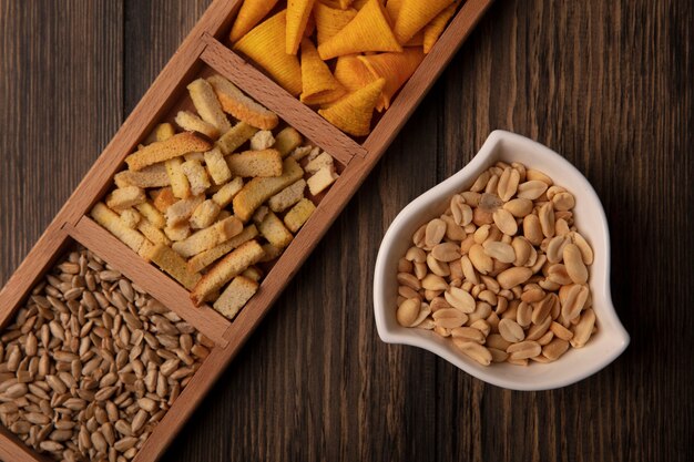 Bovenaanzicht van knapperige kegelvorm maïs snacks op een houten onderverdeelde plaat met gepelde zonnebloempitten met pijnboompitten op een kom op een houten tafel