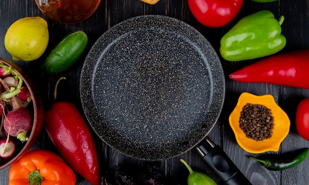 Bovenaanzicht van koekenpan en verse groenten kleurrijke paprika tomaten radijs komkommers en zwarte peperkorrels rond gerangschikt op donkere houten tafel