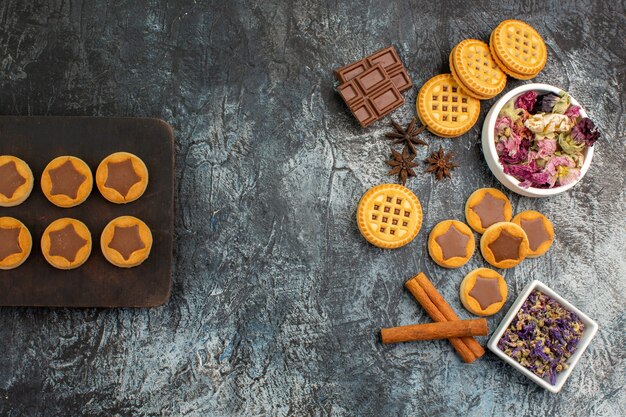 Bovenaanzicht van koekjes op houten schotel en chocoladerepen en kommen met droge bloemen op grijze grond