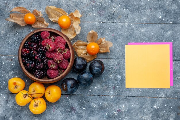 Bovenaanzicht van kom met bessen vers rijp fruit met gele kersen en pruimen op grijs, bessen fruit vers zacht bos