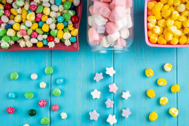 Bovenaanzicht van kommen met verschillende kleurrijke snoepjes en marshmallow verspreid uit een glazen pot op blauwe achtergrond