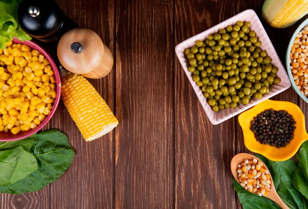 Bovenaanzicht van kommen van groene erwten maïs zaden en zwarte peper met spinazie op hout