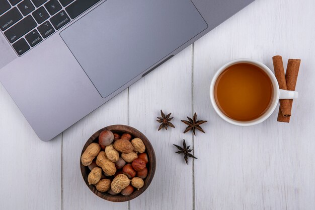 Bovenaanzicht van kopje thee met kaneel en noten in een kom en met een laptop op een wit oppervlak