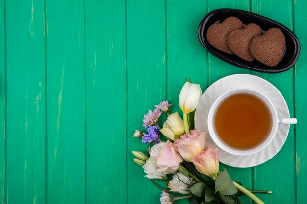 Bovenaanzicht van kopje thee op schotel en hartvormige koekjes in langwerpige kom met bloemen op groene achtergrond met kopie ruimte