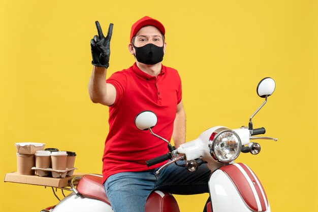 Bovenaanzicht van lachende courier man dragen rode blouse en muts handschoenen in medische masker leveren bestelling zittend op scooter overwinning gebaar maken