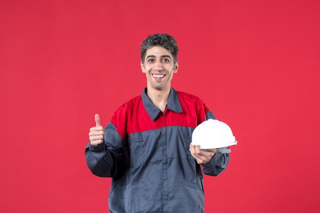 Bovenaanzicht van lachende jonge bouwer in uniform met harde hoed die een goed gebaar maakt op geïsoleerde rode muur