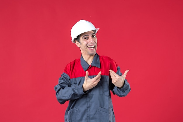 Bovenaanzicht van lachende jonge bouwer in uniform met helm die precies iets maakt op geïsoleerde rode muur