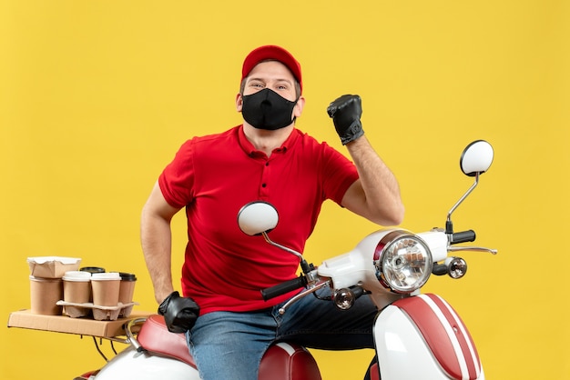 Bovenaanzicht van lachende jonge volwassene met rode blouse en hoed handschoenen in medisch masker leveren bestelling zittend op scooter terug te wijzen