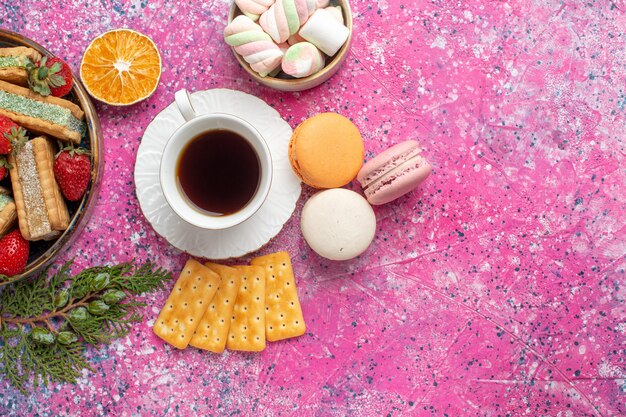 Bovenaanzicht van lekkere wafelkoekjes met franse macarons en kopje thee op roze oppervlak