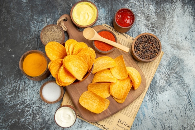 Bovenaanzicht van lekkere zelfgemaakte chips gesneden aardappelschijfjes op houten snijplank en verschillende kruiden op krant op grijze tafel