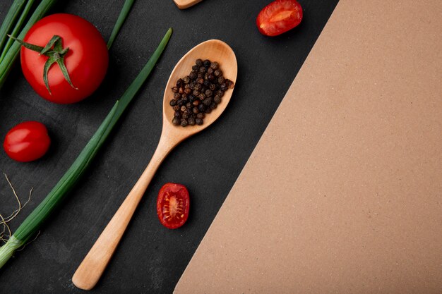 Bovenaanzicht van lepel vol peper kruiden met tomaten en lente-uitjes op zwart oppervlak met kopie ruimte