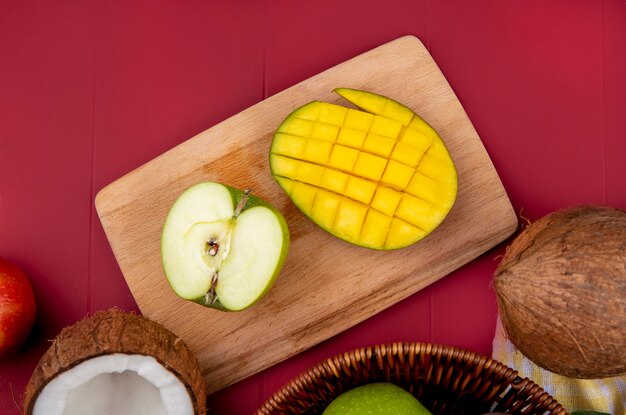 Bovenaanzicht van mango met gehalveerde groene appel op een houten keuken bord op rode ondergrond
