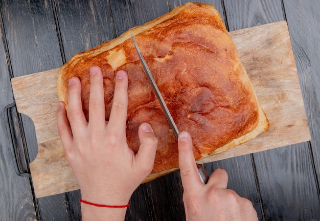 bovenaanzicht van mannelijke handen snijden zelfgebakken brood met mes op snijplank op houten achtergrond