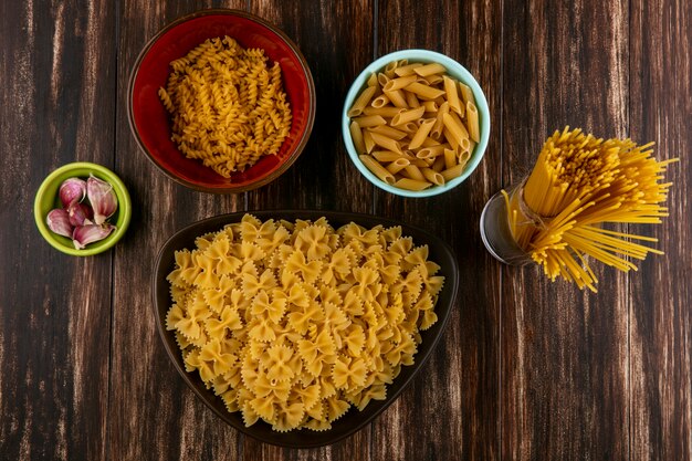 Bovenaanzicht van mix van rauwe pasta in kommen met rauwe spaghetti en knoflook op een houten oppervlak