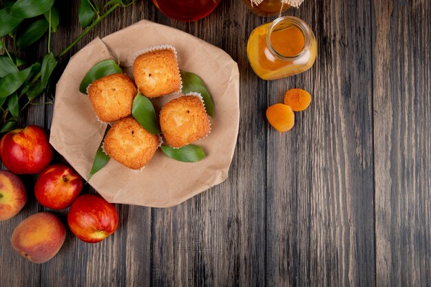 Bovenaanzicht van muffins met groene bladeren op ambachtelijk bruin papier met verse rijpe nectarines en perzikjam in een glazen pot op rustiek hout met kopie ruimte