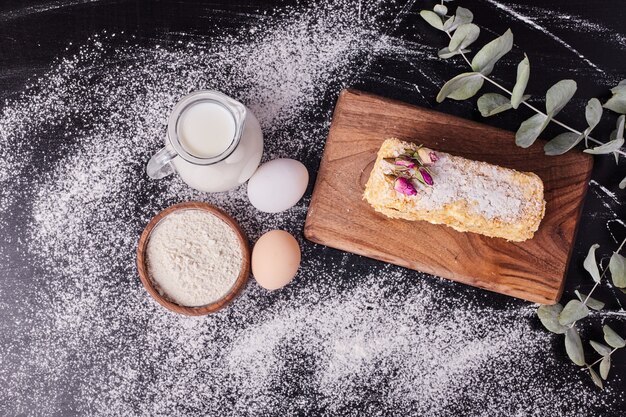 Bovenaanzicht van napoleon cake naast eieren, bloem en melk op zwarte achtergrond.