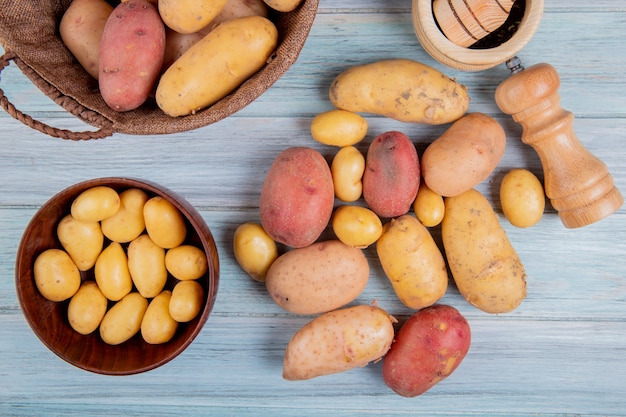 Bovenaanzicht van nieuwe aardappelen in kom en anderen van verschillende soorten in mand met knoflook crusher zout en andere aardappelen op houten oppervlak