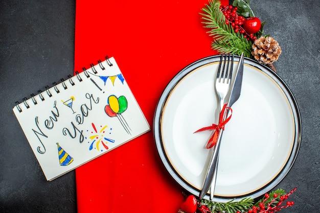 Bovenaanzicht van Nieuwjaar achtergrond met bestek met rood lint op een bord decoratie accessoires fir takken naast notebook met pen op een rood servet