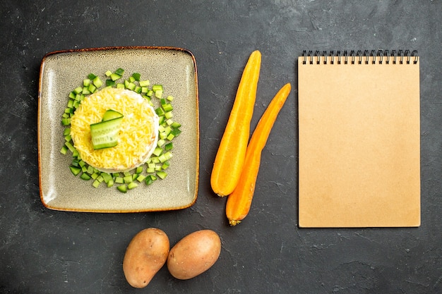 Bovenaanzicht van notitieboekje en heerlijke salade geserveerd met gehakte komkommer en wortelen met aardappelen op donkere achtergrond