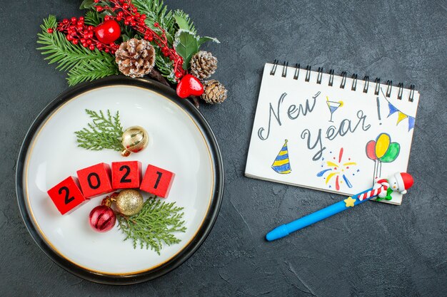 Bovenaanzicht van nummers decoratie accessoires op een plaat fir takken conifer kegel en notebook met Nieuwjaar schrijven en tekeningen op donkere achtergrond