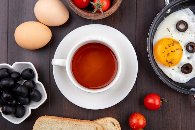 Bovenaanzicht van ontbijtset met kopje thee op schotel gebakken ei zwarte olijven brood sneetjes eieren en tomaten op hout