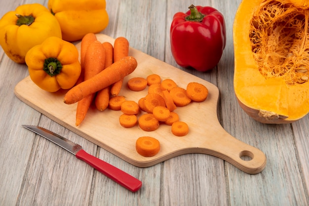 Bovenaanzicht van oranje geschilde wortelen op een houten keukenplank met mes met kleurrijke paprika geïsoleerd op een grijze houten achtergrond