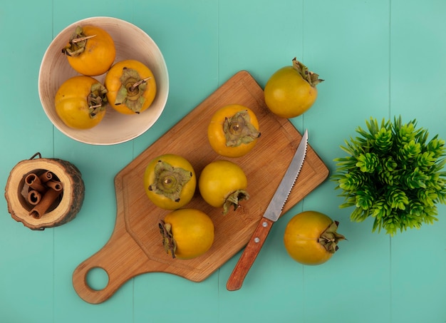 Bovenaanzicht van oranje ronde kaki vruchten op een houten keuken bord met mes kaneelstokjes op een houten pot op een blauwe houten tafel