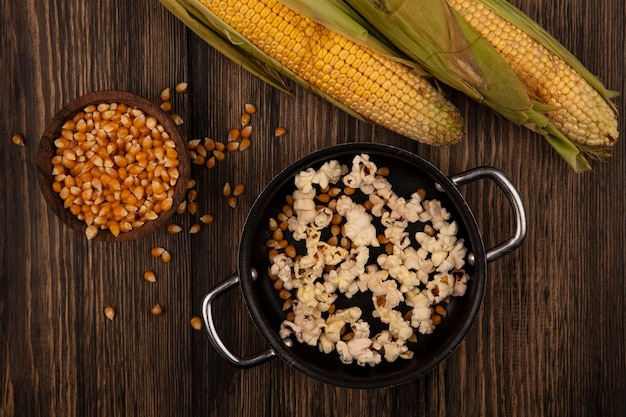 Bovenaanzicht van pan met lekkere popcorns met maïskorrels met verse likdoorns geïsoleerd op een houten kom op een houten tafel