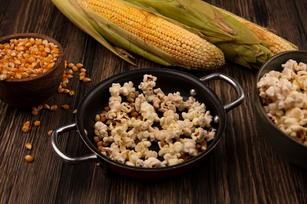 Bovenaanzicht van pan met popcorn met maïskorrels met verse likdoorns geïsoleerd op een houten kom op een houten tafel